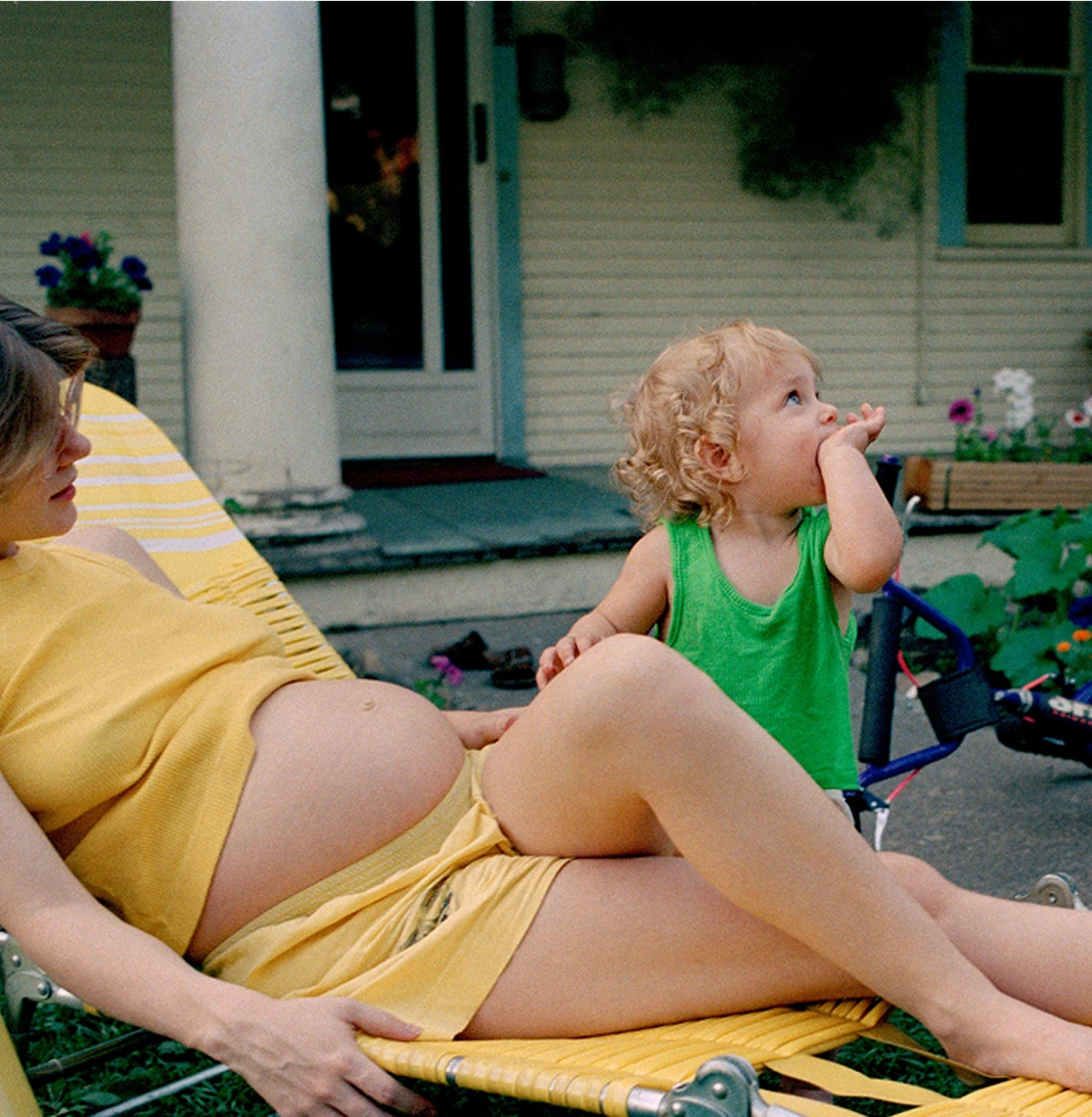 A pregnant person resting on a lawn chair while a toddler in a green tank top sits beside them in a summer yard