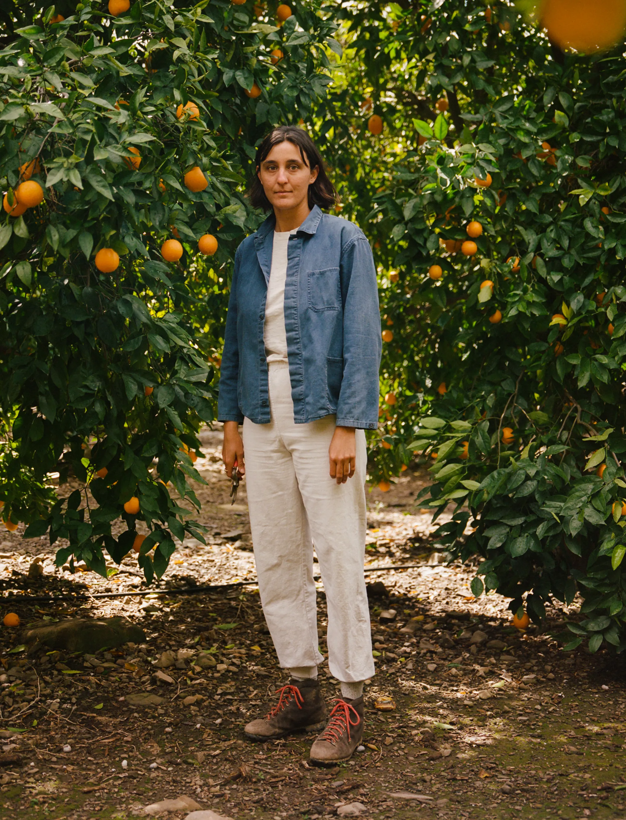 A person in a denim shirt standing in front of a leafy tree, looking calmly at the camera