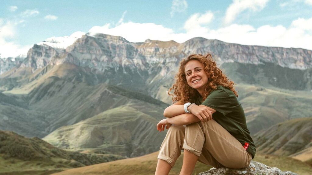 A smiling woman with red curly hair sits on a rock atop a mountain.