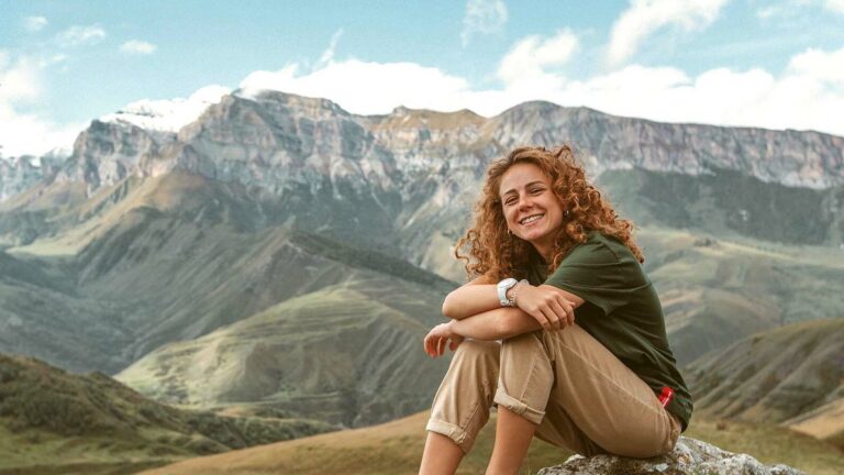 A smiling woman with red curly hair sits on a rock atop a mountain.