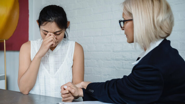 A blonde woman with a bob wearing a black blazer holds the hand of a woman sitting across from her in an attempt to comfort her as she cries.