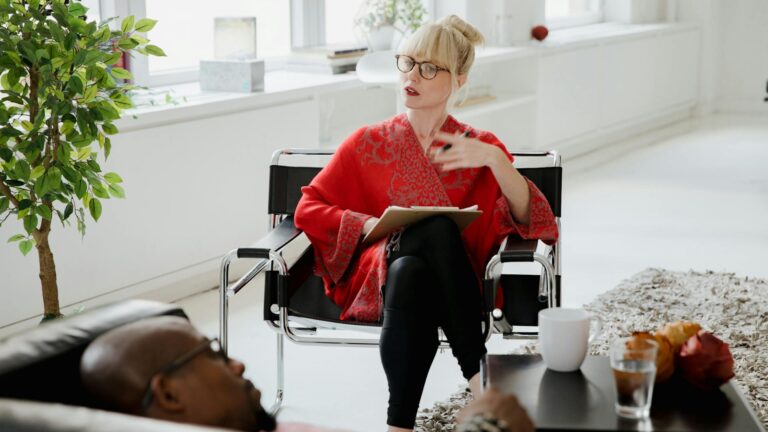 A therapist with blonde hair in a high bun wearing a red shirt sits across from a man lying on a couch and listens to him speak.