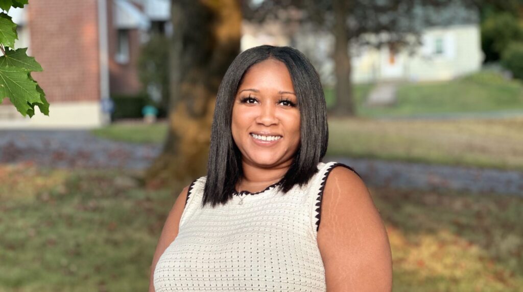 A black woman with a laptop in her lap, holding a coffee in her had as she sits on a park bench, working remotely.