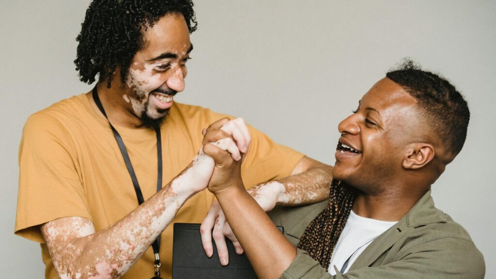 Two black men, one with vitiligo, clasp their hands together in a celebratory manner.