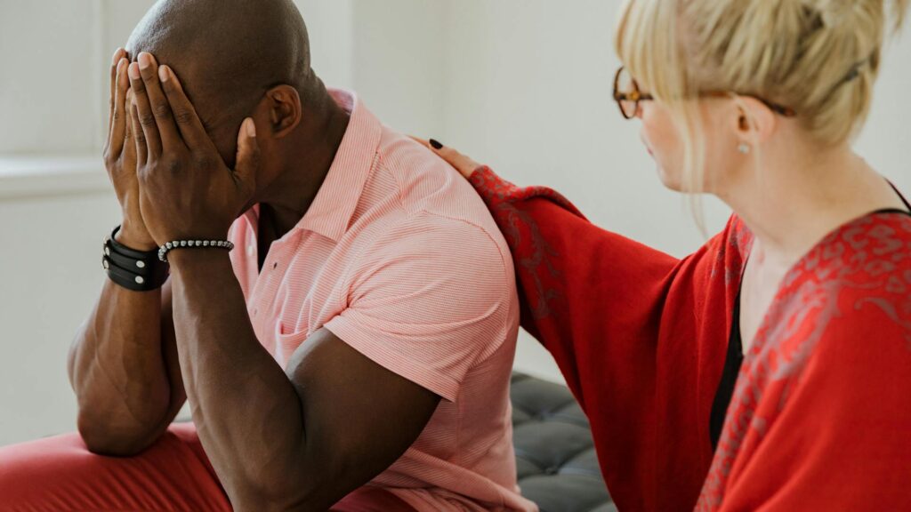 A white woman in a red shirt places a comforting arm on the shoulder of a black man in an orange shirt. The man is covering his face with his hands in a sign of distress.