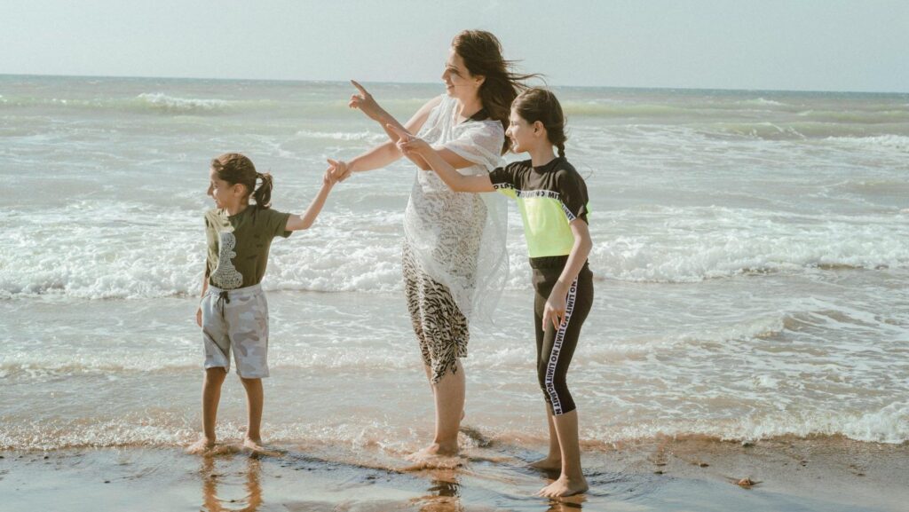 A woman in a flowy white dress stands on the shoreline of a beach with her two children.