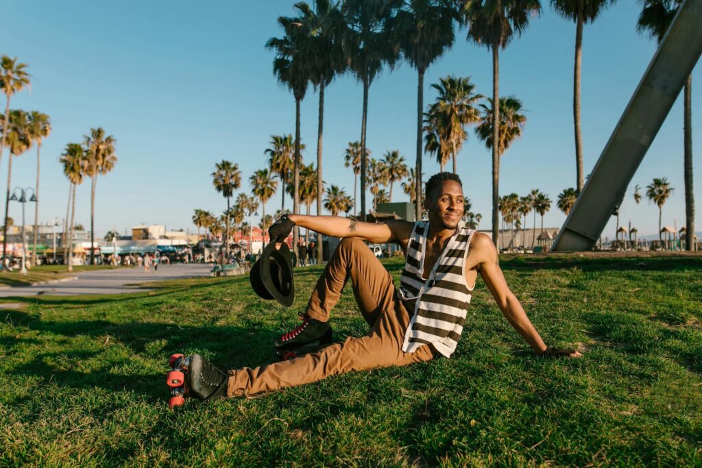A Black man wearing black roller skates sits in the grass wearing a blue and white striped shirt with palm trees in the background.