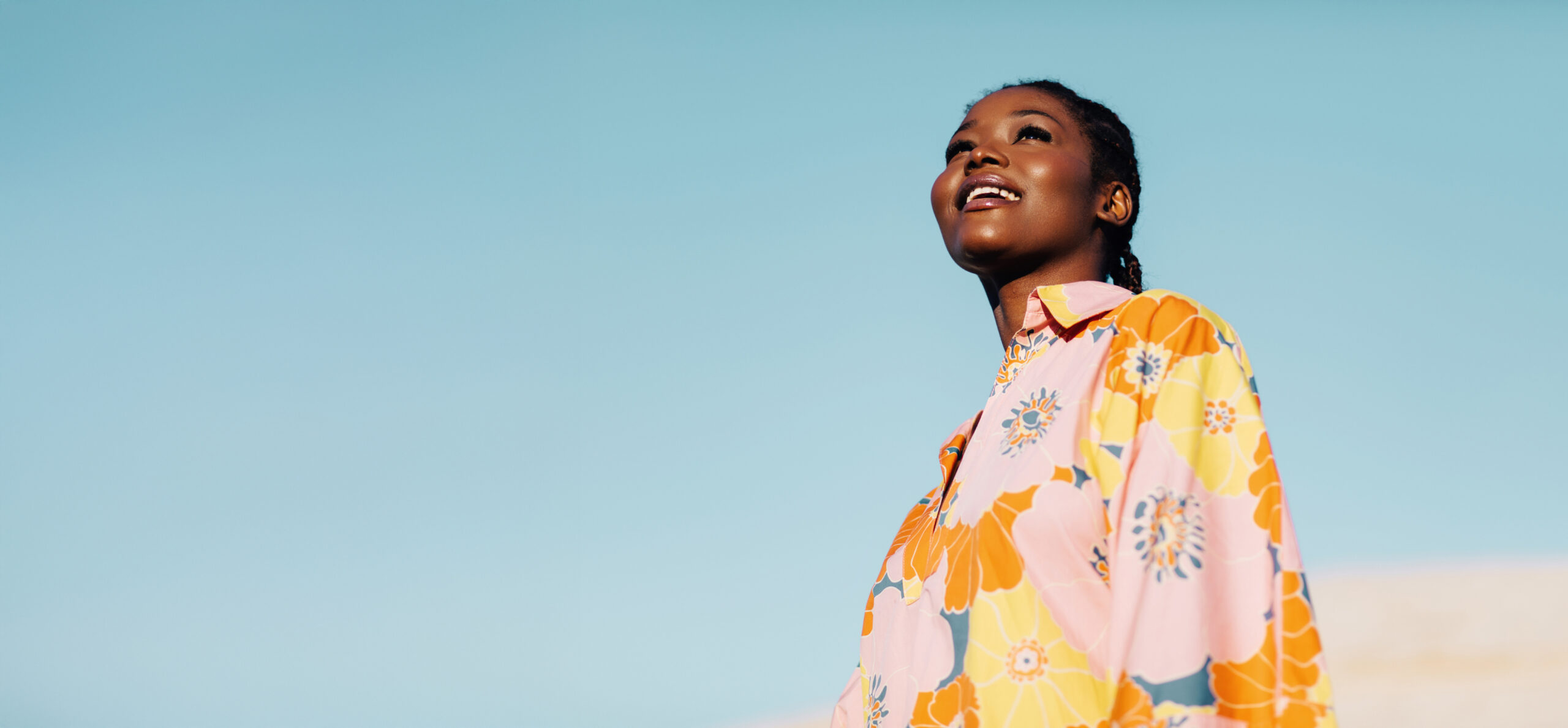 A woman smiling and looking up at the sky, wearing a colorful floral shirt against a clear blue background.