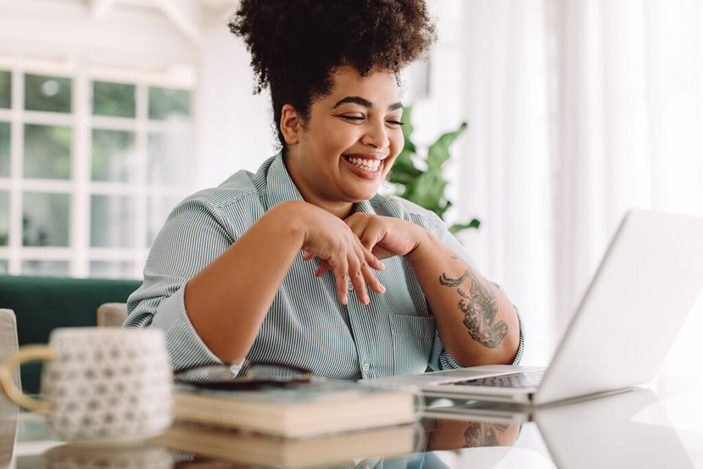 A woman wearing headphones, smiling at the laptop while she joins an online therapy session.