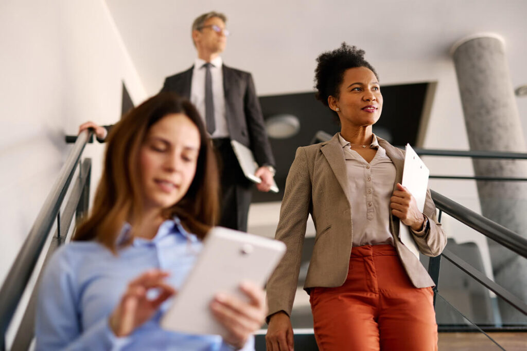 Three people walk down the stairs in an office environment. Two people carry documents while one reviews a tablet.