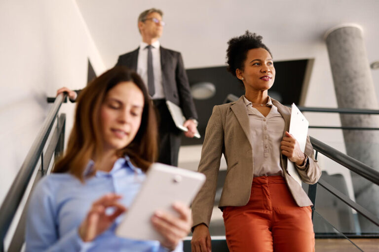 Three people walk down the stairs in an office environment. Two people carry documents while one reviews a tablet.