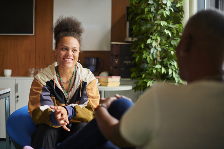 A person smiles with clasped hands in conversation with a therapist who is in the foreground facing away.