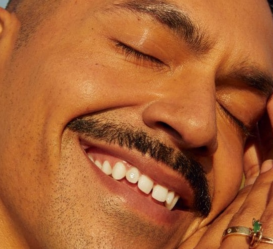 A person with a mustache smiles blissfully with eyes closed, face bathed in warm sunlight. They gently cradle their cheek, conveying happiness and serenity.