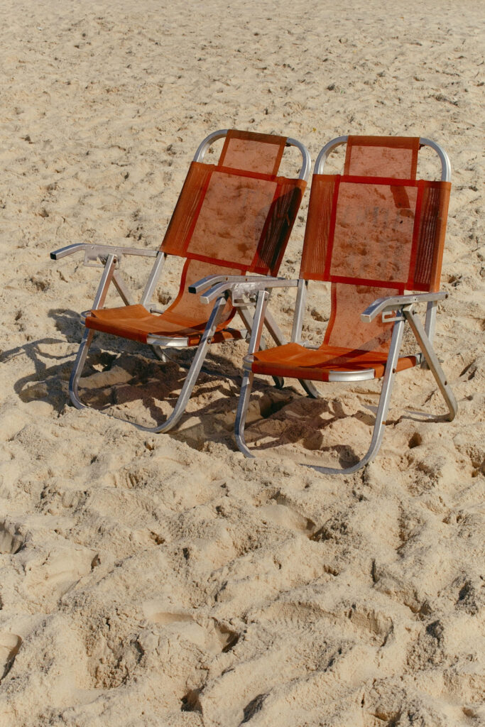 Two orange beach chairs sit empty on sunlit sand, evoking a sense of relaxation and leisure. Shadows cast softly, enhancing the serene beach atmosphere.