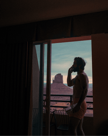 A man stands in a dimly lit room, looking out a glass door towards a scenic desert landscape at sunset, conveying a contemplative and serene mood.