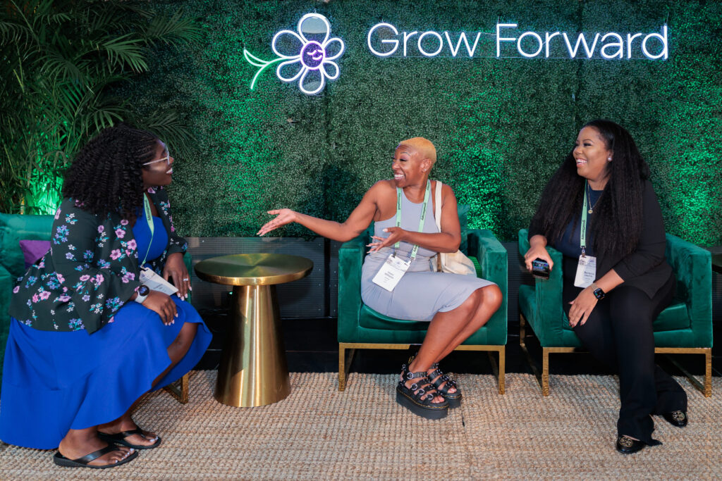 Three women sit on green chairs, laughing and chatting, in front of a plant wall with a "Grow Forward" sign. The atmosphere is joyful and relaxed.