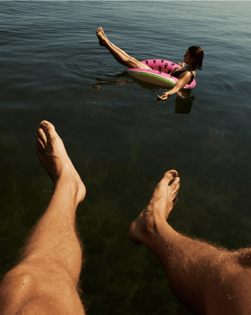 Two people in a lake, one relaxing on a pink donut-shaped float, the other sitting with just legs visible, creating a serene and carefree vibe.
