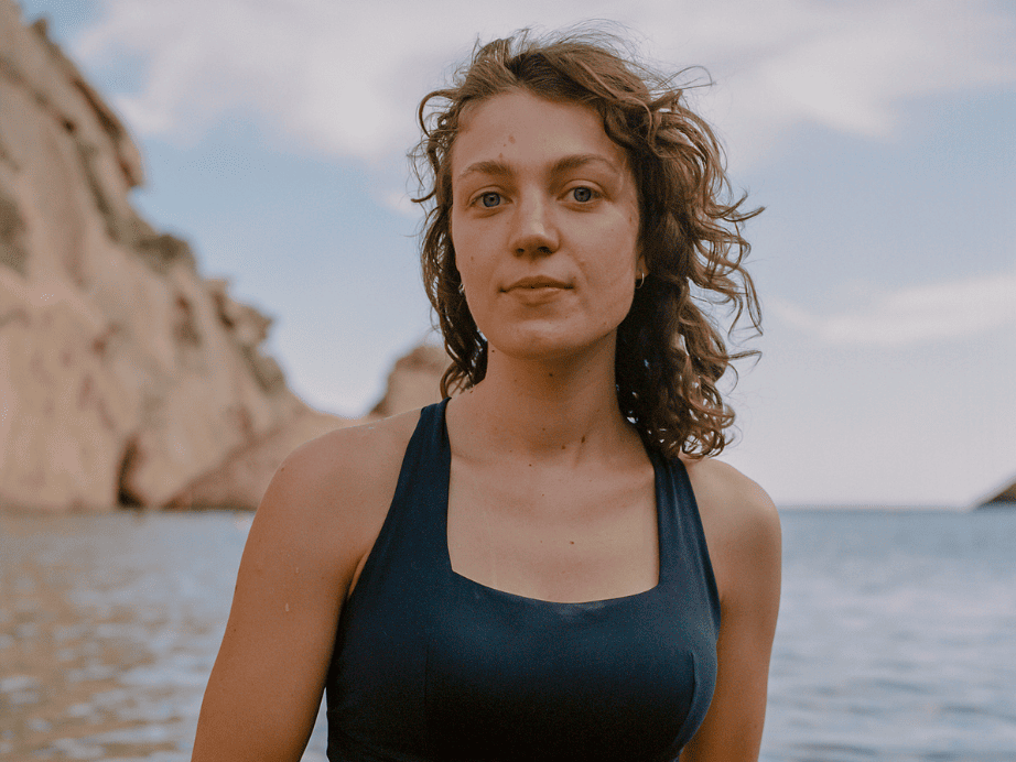Young woman in a navy swimsuit stands waist-deep in calm ocean water, with rocky cliffs in the background. She gazes confidently at the camera.