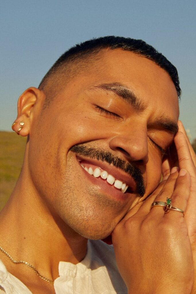 A person with short hair and a mustache smiles joyfully with closed eyes, basking in sunlight. They wear earrings, rings, and a chain, creating a serene, content mood.