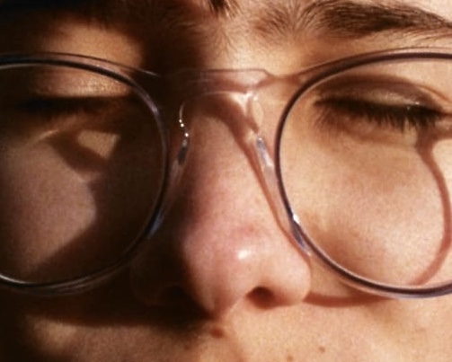 Close-up of a person with eyes closed, wearing round glasses. Sunlight casts a shadow on their face, creating a calm and serene mood.
