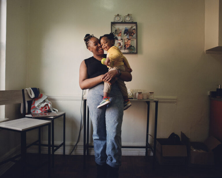 A woman lovingly holds a smiling child in a cozy room with soft lighting. The child wears a yellow outfit, evoking warmth and happiness.