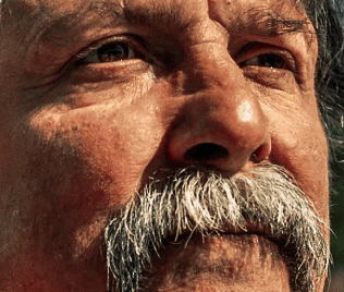 Close-up of an older man's face with a thick gray mustache, wrinkled skin, and visible sunlight reflecting in his eyes.