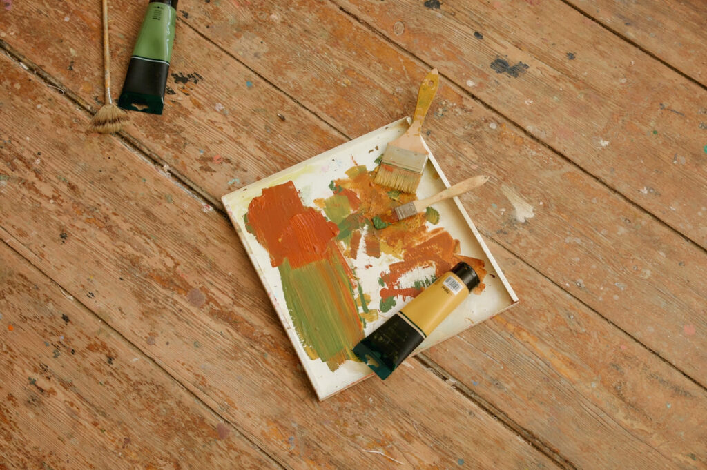 A paint tray with mixed orange and green colors and used brushes lies on a rustic wooden floor, evoking a creative, artistic atmosphere.
