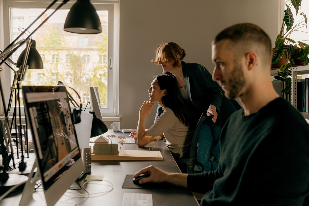 A team collaborates in an office with natural light. A man works on a computer, while two women discuss a model. The mood is focused and productive.