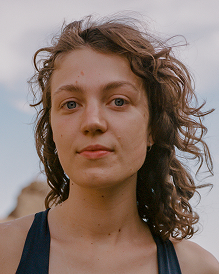 A woman with wavy hair gazes into the camera with a calm expression. She's outdoors, under a blue sky with clouds. She wears a black top, conveying a serene mood.
