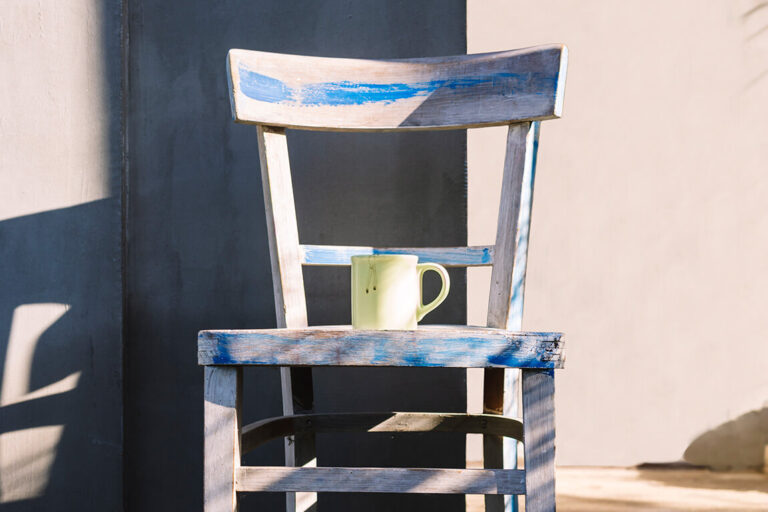 A light yellow coffee mug sitting on a paint-stripped wooden chair with cement walls in the background.